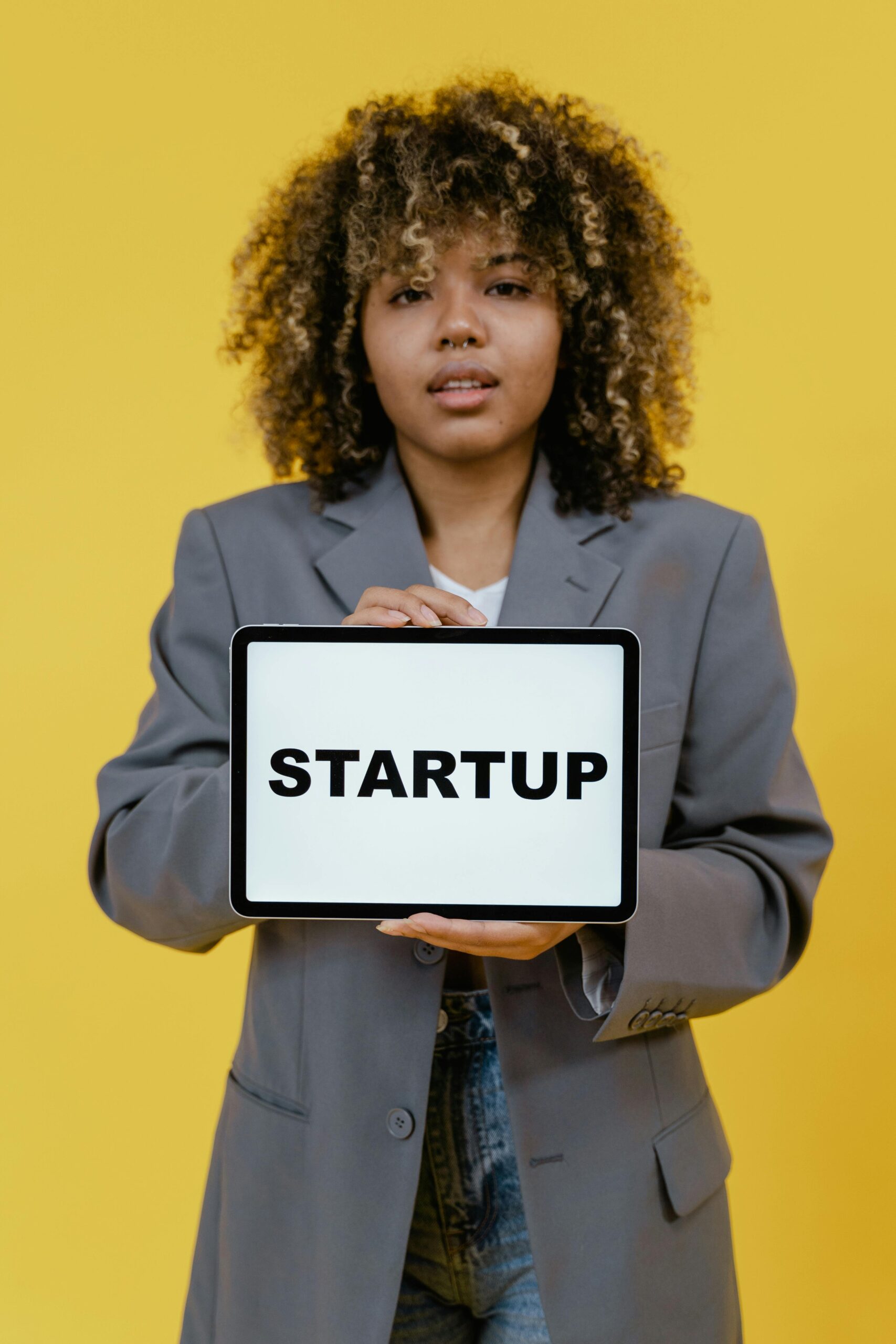 A young woman with curly hair holds a tablet displaying 'Startup' against a yellow background.